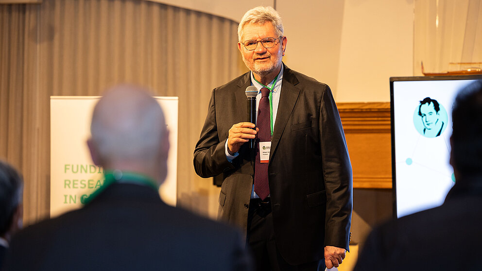 Robert Schlögl, der Präsident der Humboldt-Stiftung steht auf einer Bühne und spricht mit einem Mikrophon zum Publikum. Im Hintergrund sieht man das Logo der Humboldt-Stiftung.