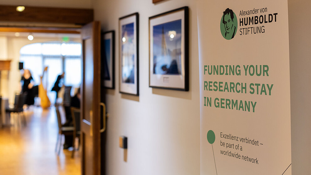 Ein Schild mit der Aufschrift "Funding your research stay in Germany. Exzellenz verbindet - be part of a worldwide network" steht neben dem Eingang zu einem Raum. Man kann in den Raum hineinsehen.