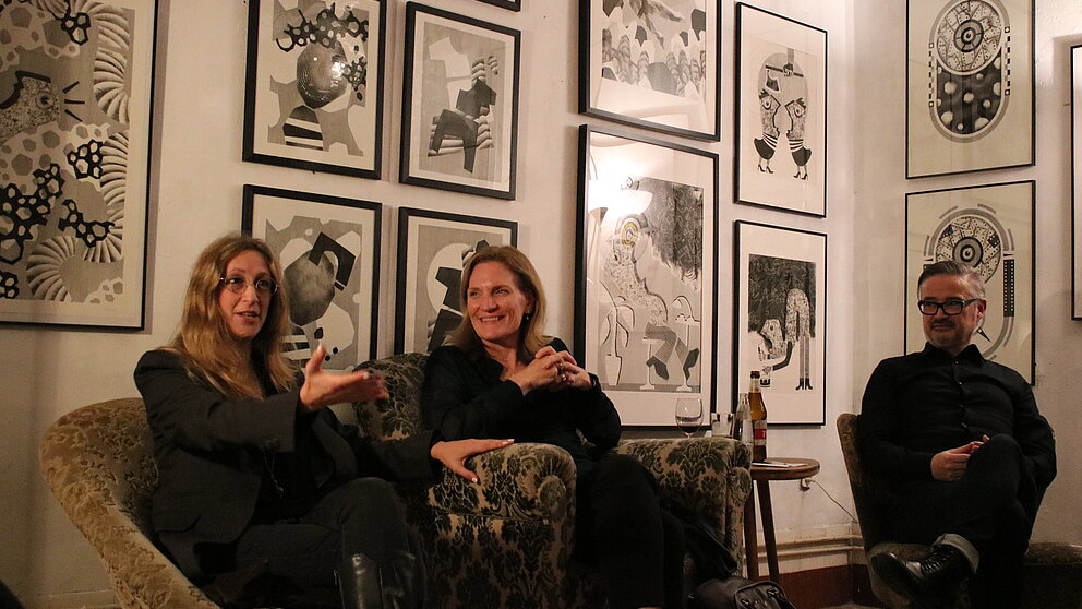 Three people sit on armchairs in a room with many framed black and white pictures on the wall.