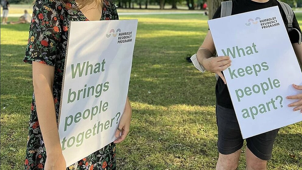 Two signs held by two people in a park with inscriptions:  “What brings people together?” and “What keeps them apart?” 