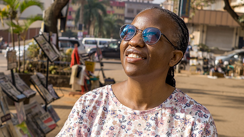 Eine Frau mit Sonnenbrille steht auf einem Platz und blickt in die Sonne, hinter ihr ein Stand mit Zeitungen und Zeitschriften