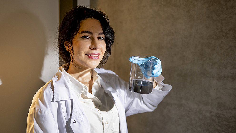 Eine lächelnde Frau in einem Laborkittel hält ein Becherglas mit einer dunklen Flüssigkeit in der Hand.