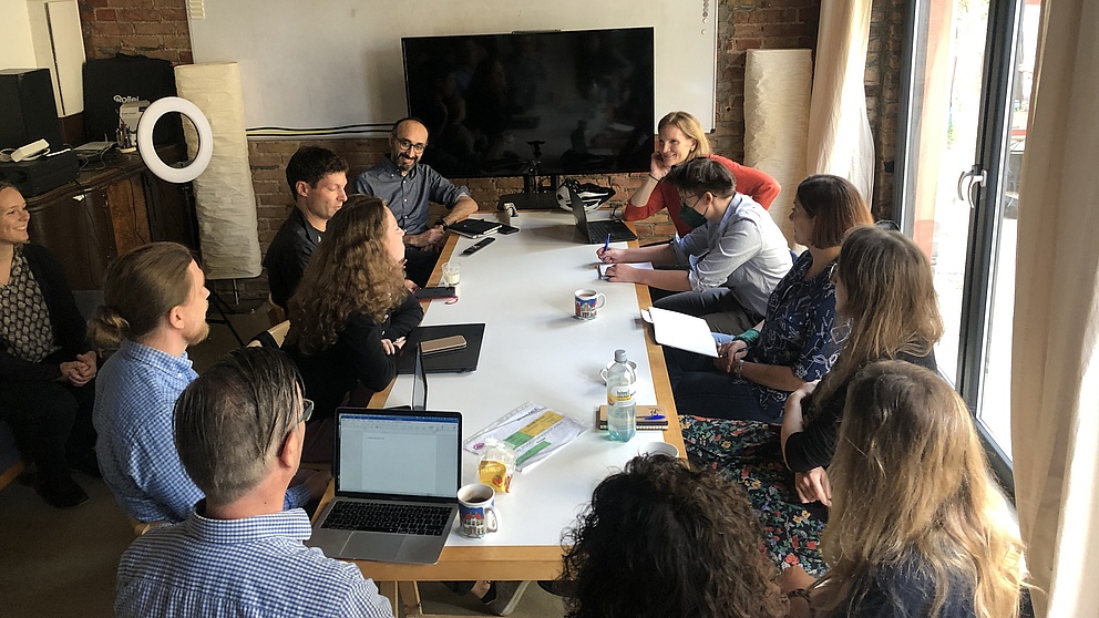 12 Personen sitzen um einen Tisch in einem Zimmer mit Laptops und Notizblöcken und diskutieren