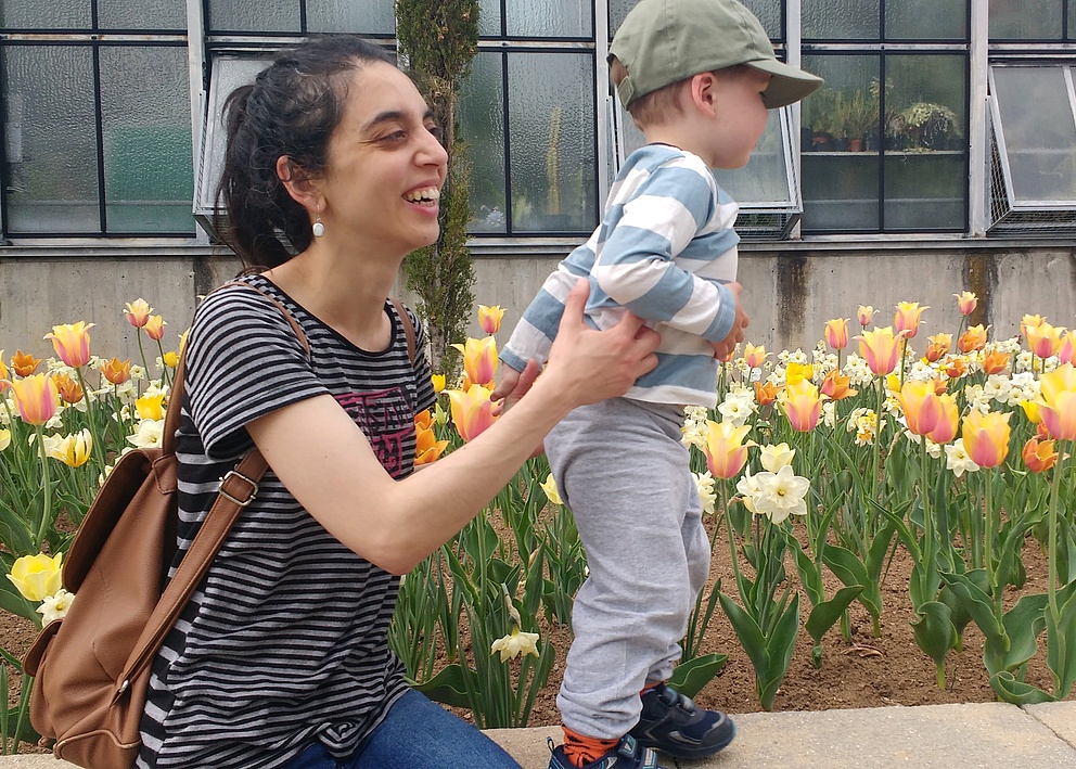 Das Foto zeigt Paula Tribelli, wie sie ihr Kleinkind hält. Beide tragen gestreifte Oberteile und lächeln glücklich. Hinter ihnen befindet sich ein Beet mit Tulpen.