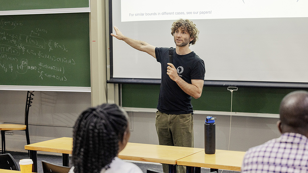 Ein Dozent mit lockigem Haar präsentiert mit einem Mikrofon in der Hand etwas vor einer Gruppe von Zuhörenden - im Hintergrund ist eine Tafel mit mathematischen Gleichungen zu sehen. 
