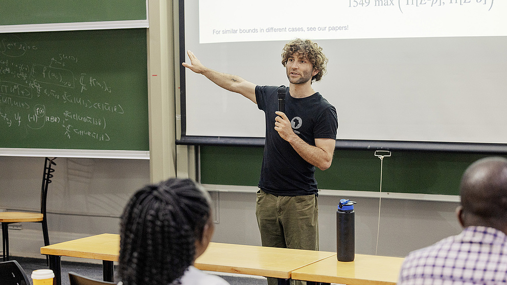 Ein Dozent mit lockigem Haar präsentiert mit einem Mikrofon in der Hand etwas vor einer Gruppe von Zuhörenden - im Hintergrund ist eine Tafel mit mathematischen Gleichungen zu sehen. 