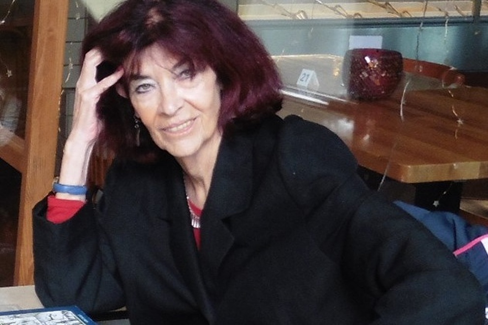 Eine Frau in einem schwarzen Mantel sitzt mit Blick in die Kamera an einem Tisch, auf dem ein Buch und eine Brille liegen