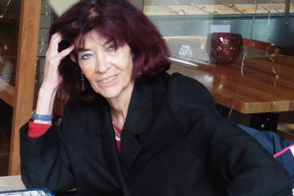 Eine Frau in einem schwarzen Mantel sitzt mit Blick in die Kamera an einem Tisch, auf dem ein Buch und eine Brille liegen