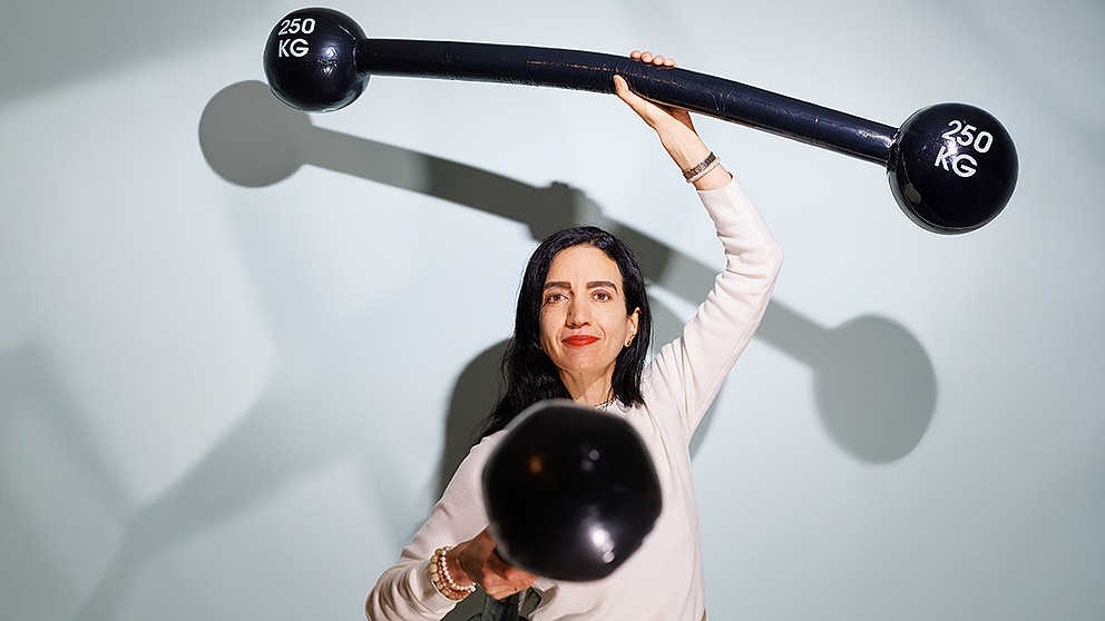 Eine Frau, die zwei Gewichte hochhält, schaut freundlich-bestimmt in die Kamera – ihre Gewichte symbolisieren ihr wissenschaftliches Kernthema „Kinder stärken“.