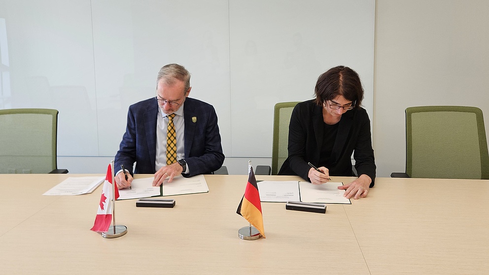 Zwei Menschen sitzen hinter einem Tisch und unterschreiben etwas. Vor ihnen stehen die kanadische und die deutsche Flagge auf einem Tisch.
