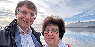 Martina Havenith und Albert Newen stehen vor einem See mit Bergen im Hintergrund. Albert Newen trägt ein Hemd mit schwarzer Lederjacke, Martina Havenith eine weiße Bluse mit bunter Strickjacke.