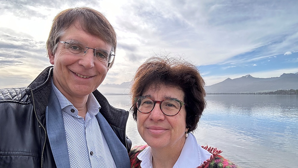 Martina Havenith and Albert Newen standing in front of a lake with mountains in the background. Albert Newen is wearing a shirt with a black leather jacket, while Martina Havenith is dressed in a white blouse with a colorful knitted jacket.