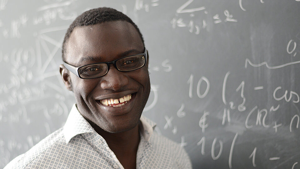 Mouhamed Moustapha Fall: ehemaliger Humboldt-Forschungsstipendiat und Inhaber des deutschen Forschungslehrstuhls am AIMS in Senegal vor einer Tafel mit mathematischen Formeln