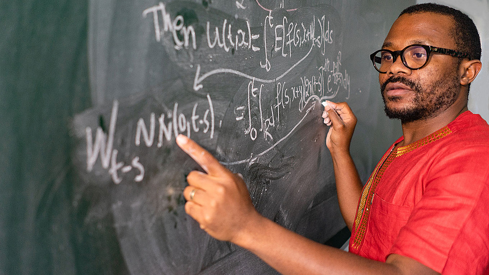 Olivier Pamen vor einer Tafel mit mathematischen Formeln mit Link zum Dossier