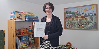 Eine Frau in einem eleganten Outfit zeigt ein Zertifikat, während im Hintergrund ein farbenfrohes Wandbild und Spielzeugregale zu sehen sind.