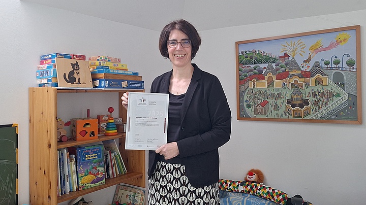 Eine Frau in einem eleganten Outfit zeigt ein Zertifikat, während im Hintergrund ein farbenfrohes Wandbild und Spielzeugregale zu sehen sind.