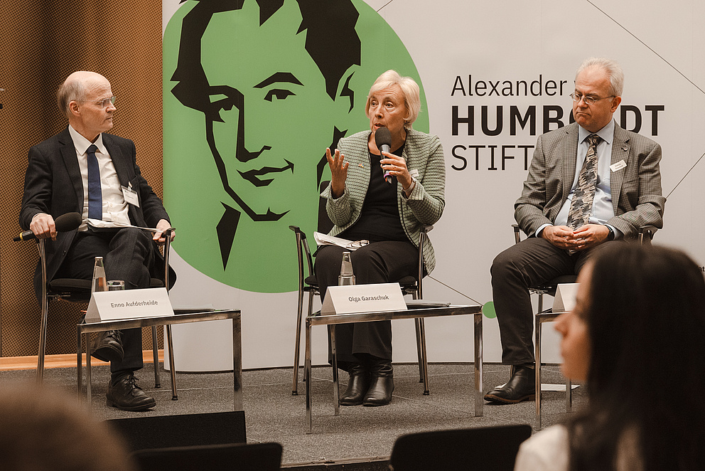 Two men and a woman on the podium, the woman in the middle speaks into a microphone.