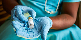 Stockfoto: Eine Person in Laborkleidung hält ein Fläschchen, auf dem COVID-19 Vaccine steht. 