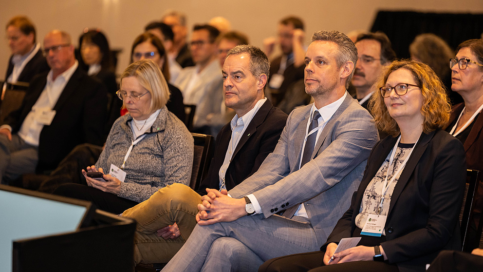 Das Publikum wird von vorne gezeigt. Es hört einer Rede zu.