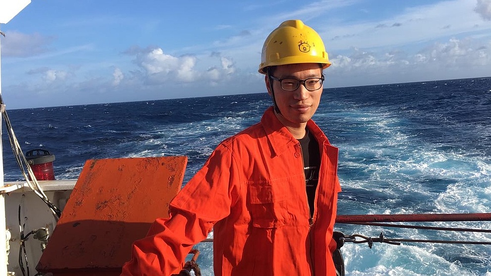 Ein Mann in orangefarbener Arbeitskleidung und gelbem Helm steht auf einem Schiff, während Wellen hinter ihm aufschäumen.