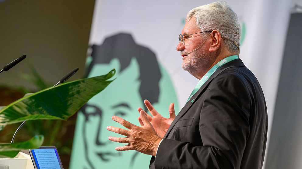 Ein Mann hält eine Rede hinter einem Podium mit dem Logo der Humboldt-Stiftung im Hintergrund und verwendet Gesten.