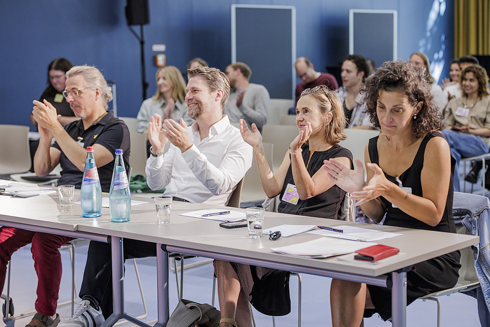 Vier Menschen sitzen hinter einem Tisch, lächeln und klatschen.