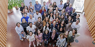 Eine Gruppe von Menschen steht in einem Raum und wird von oben fotografiert. Alle gucken in die Kamera und lächeln.