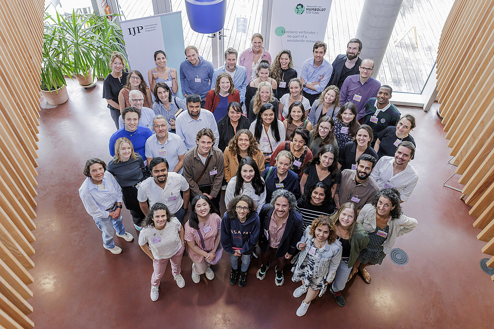 Eine Gruppe von Menschen steht in einem Raum und wird von oben fotografiert. Alle gucken in die Kamera und lächeln.