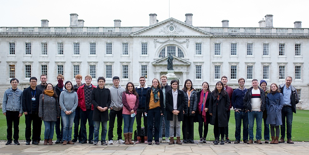 Das Foto zeigt Silvia Vignolini mit über 20 Studierenden, die lächelnd vor einem Gebäude der Universität Cambridge auf einem grünen Rasen stehen. Das Gebäude zeichnet sich durch seine klassische Architektur und eine Skulptur in der Mitte aus.