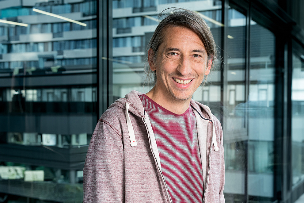 Portraitbild eines lachenden Mannes mit braunen Haaren und einer Kapuzenjacke. 