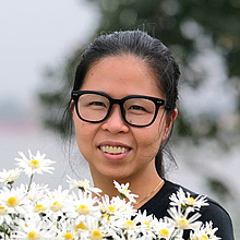 Foto einer Frau mit schwarzen Haaren und Brille, im Vordergrund weiße Blumen