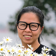 Foto einer Frau mit schwarzen Haaren und Brille, im Vordergrund weiße Blumen