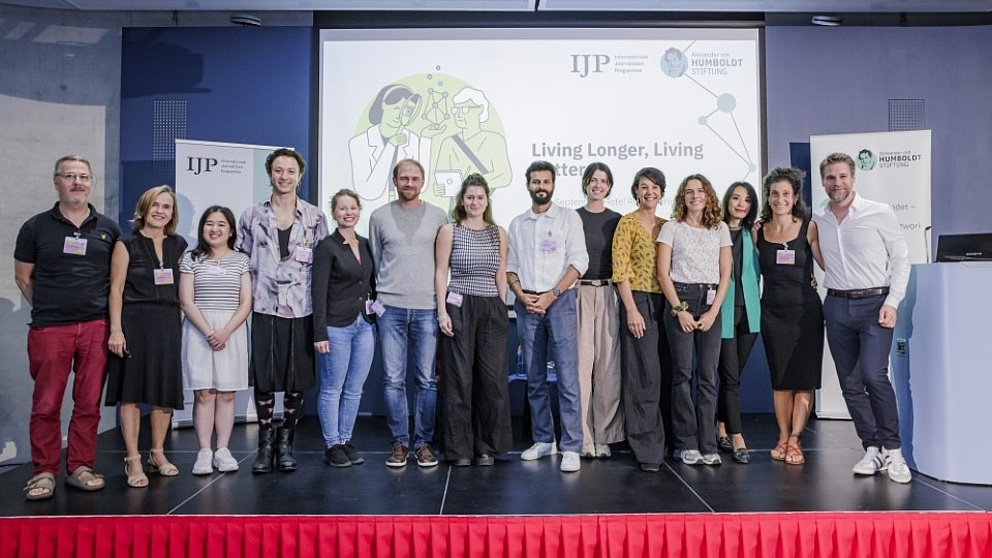 Eine Gruppe von zwölf Personen steht auf einer Bühne, lächelnd, vor einem Bildschirm mit dem Titel 'Living longer, Living better'.