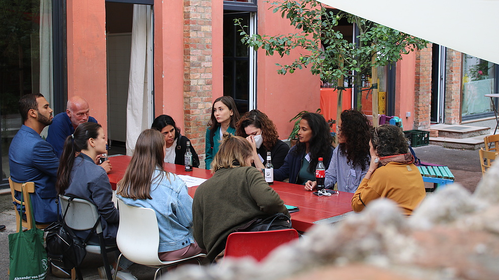 Elf Personen sitzen an einem Tisch in einem Garten und diskutieren