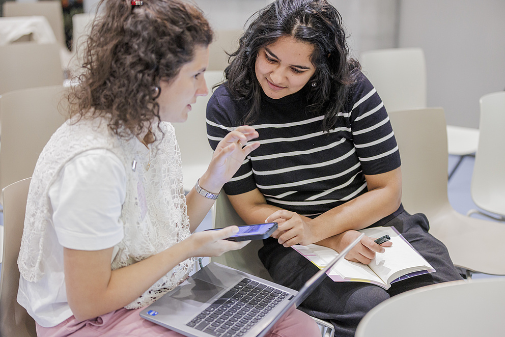 Zwei Frauen sitzen nebeneinander auf Stühlen. Eine Frau zeigt etwas auf ihrem Handy, die andere schaut darauf.
