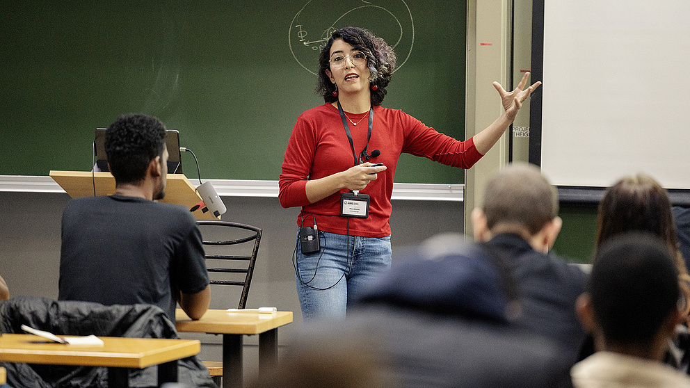 Eine Dozentin in einem roten Oberteil erklärt lebhaft ein Thema vor einer Gruppe von Studierenden in einem Klassenzimmer.