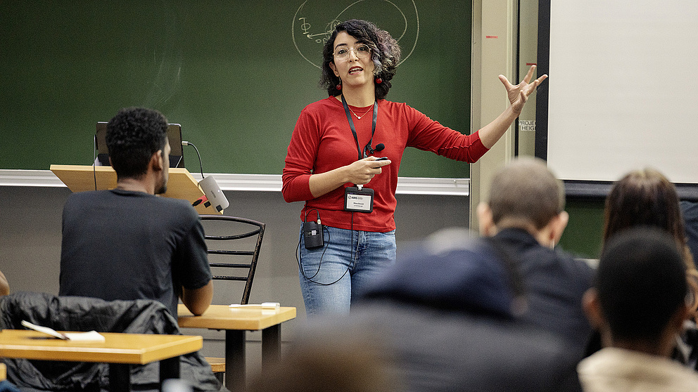 Eine Dozentin in einem roten Oberteil erklärt lebhaft ein Thema vor einer Gruppe von Studierenden in einem Klassenzimmer.
