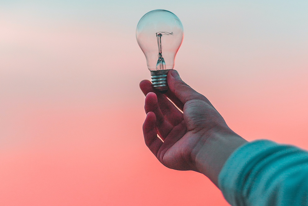 Eine Person vor einem orange-türkisen Hintergrund hält eine Glühbirne in der Hand. Es ist nur ihr Arm zu sehen.