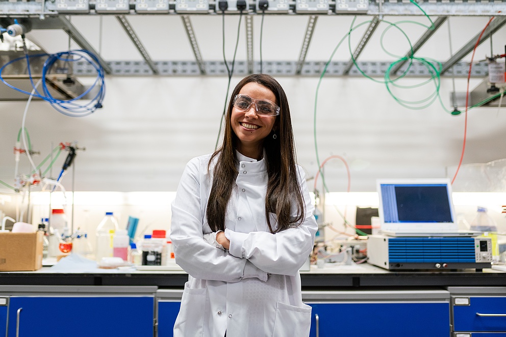 A young woman with a happy smile and her arms crossed stands confidently in a laboratory. She is wearing a white lab coat and has long, dark brown hair.