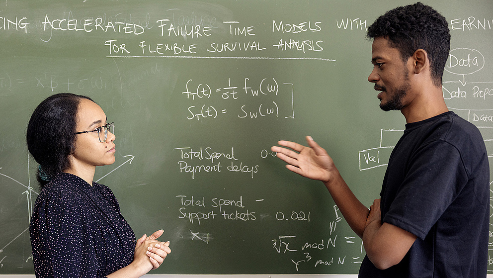 Im Vordergrund diskutieren zwei Personen an einer Tafel, die mit mathematischen Formeln und Diagrammen beschriftet ist.