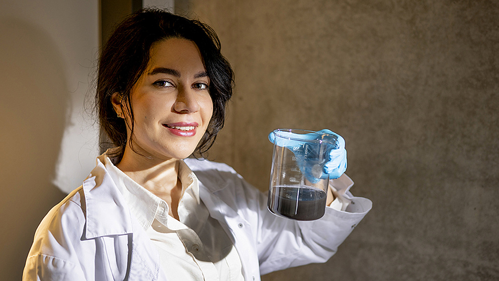 Eine lächelnde Frau in einem Laborkittel hält ein Becherglas mit einer dunklen Flüssigkeit in der Hand.