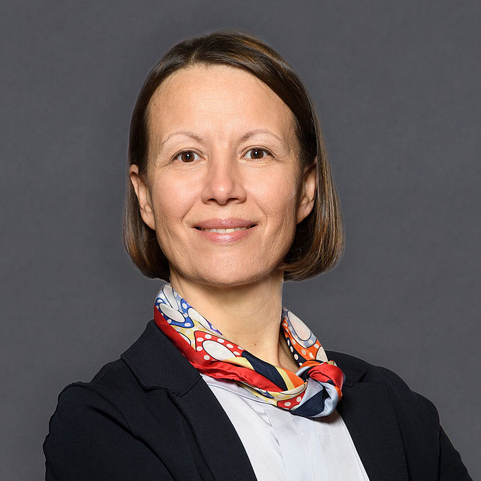 Eine Frau mit schulterlangen Haaren lächelt freundlich, während sie einen eleganten schwarzen Blazer und einen bunten Schal trägt.