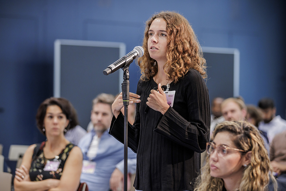 Eine Frau aus dem Publikum steht hinter einem Mikro und spricht etwas hinein. 