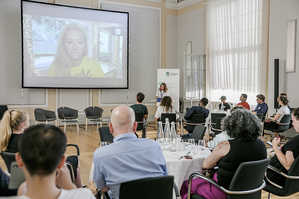 Zu sehen ist eine Menschenmenge auf Stühlen von hinten, die auf einen Bildschirm schauen. Auf dem Bildschirm ist eine Frau zu sehen, die einen Online-Vortrag hält. 
