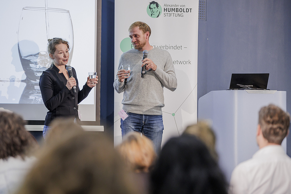 Eine Frau und ein Mann stehen auf einer Bühne, die Frau spricht in ein Mikro zum Publikum. Sie halten Wassergläser in den Händen.