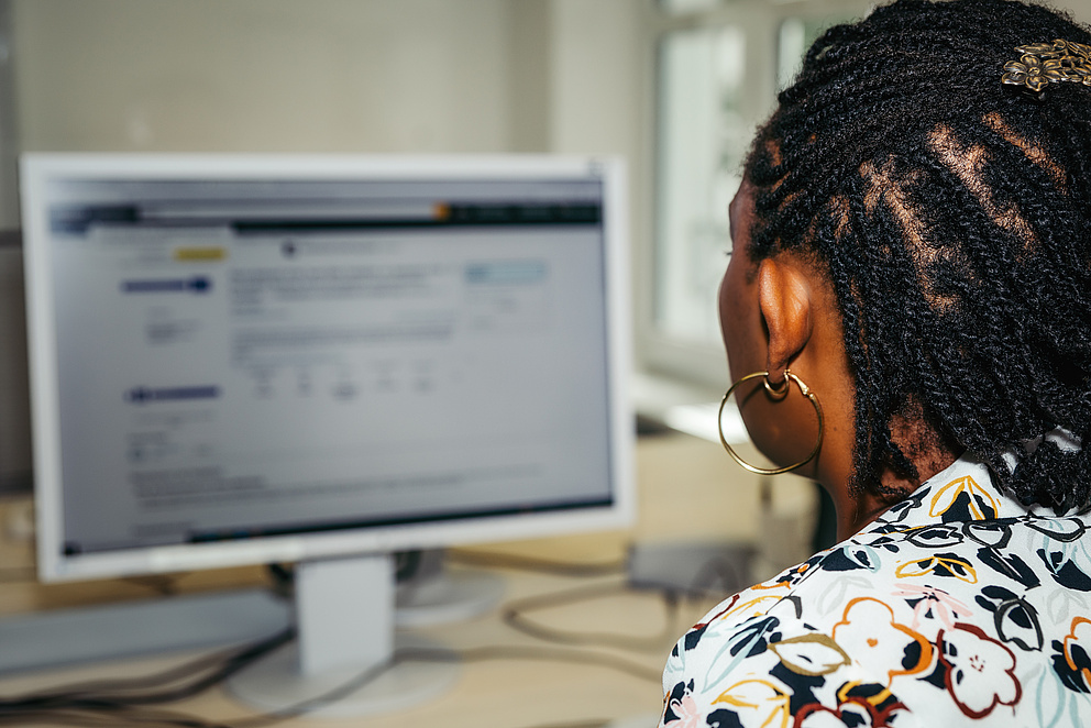 Der Hinterkopf einer Schwarzen Frau mit Braids, Kreolen und einer gemusterten Bluse ist im Fokus. Sie betrachtet einen Computer-Bildschirm, auf dem eine Website zu sehen ist. 