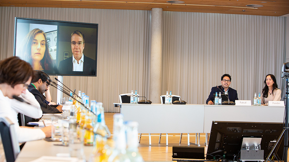 Teilnehmende eines Panels sitzen an ihren Tischen, zwei Teilnehmende sind per Video zugeschaltet