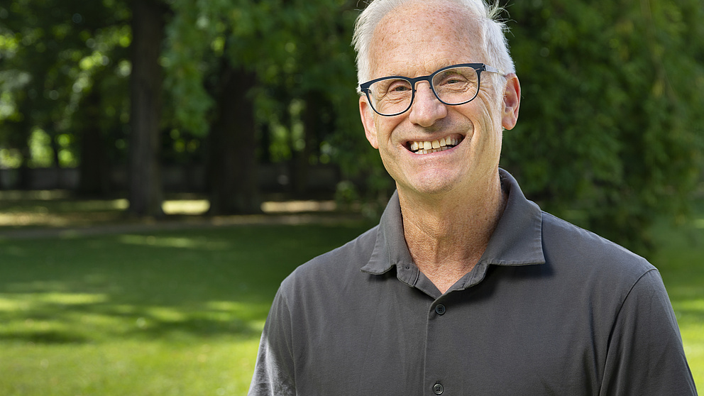 Ein lächelnder Mann mit grauen Haaren und Brille steht in einem sonnigen Park, umgeben von grünen Bäumen und Gras.