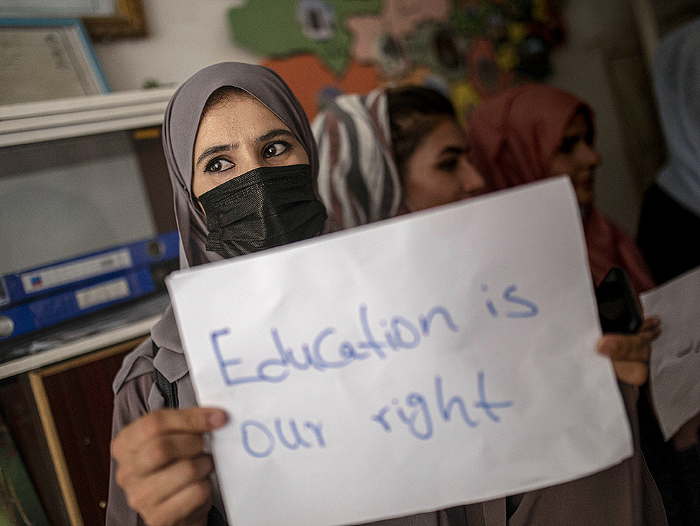 Eine junge Frau mit Kopftuch und Maske hält ein Schild mit der Aufschrift 'Bildung ist unser Recht' in einer belebten Umgebung.