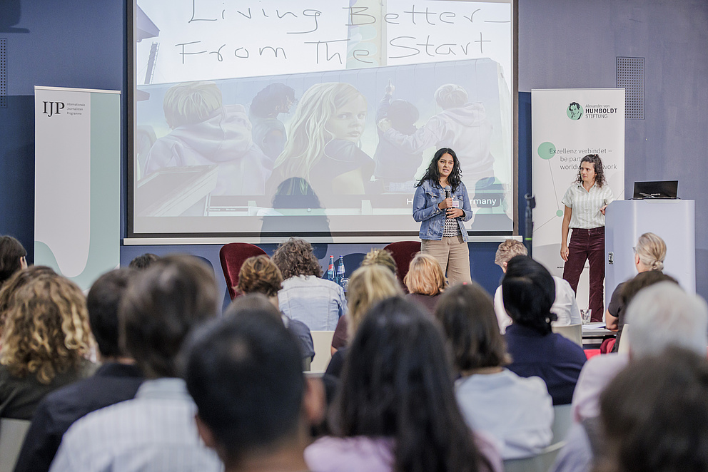 Man sieht das Publikum in einem Raum von hinten. Auf der Bühne stehen zwei Frauen, die eine spricht zum Publikum. Im Hintergrund sieht man eine Präsentation mit einem Bild eines kleinen blonden Mädchens, das sich zur Kamera umdreht und ernst hinein schaut. Text auf dem Bild: "Living Better from the start"